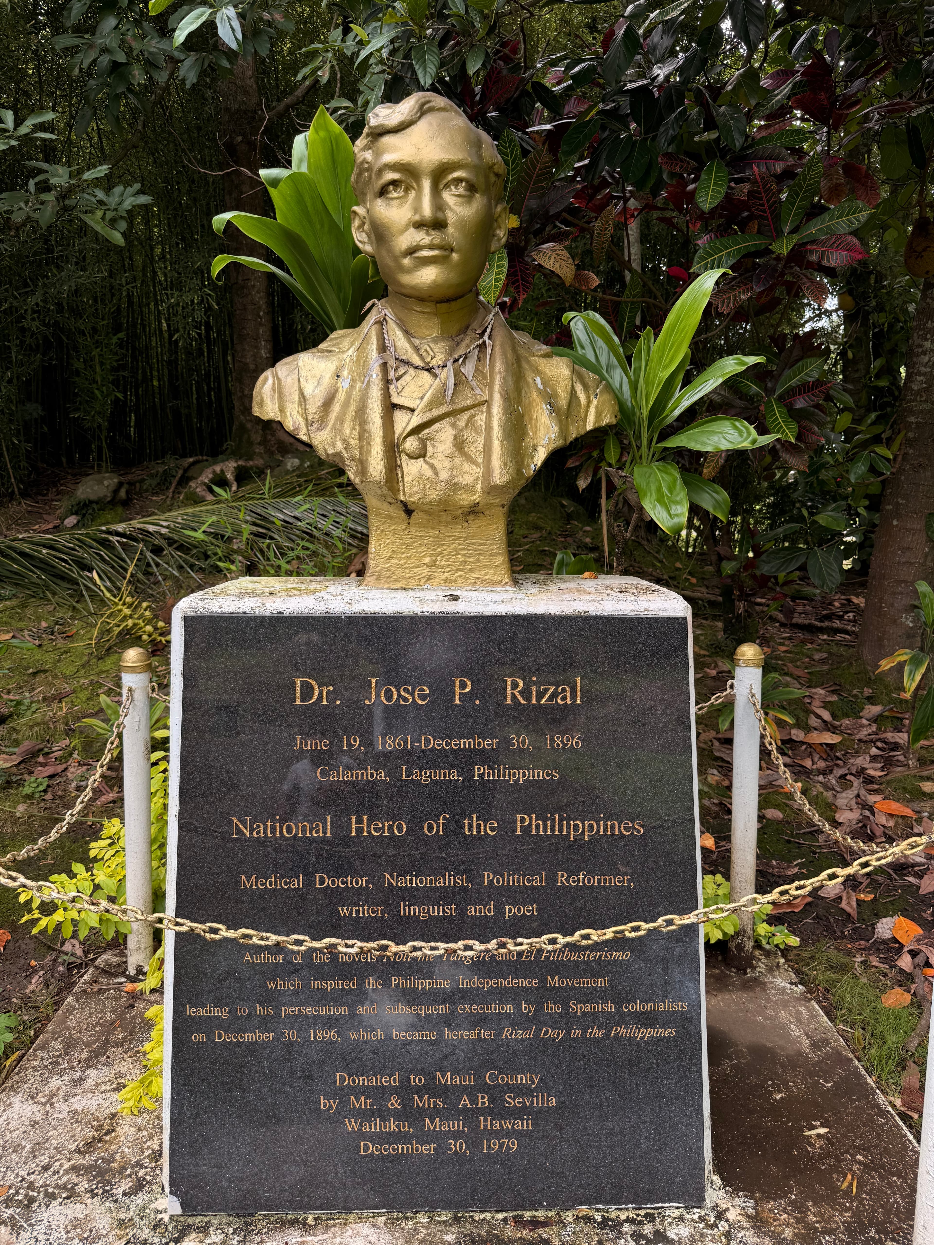Gold bust of Dr. José P. Rizal on a stone pedestal with an informational plaque, set among tropical foliage