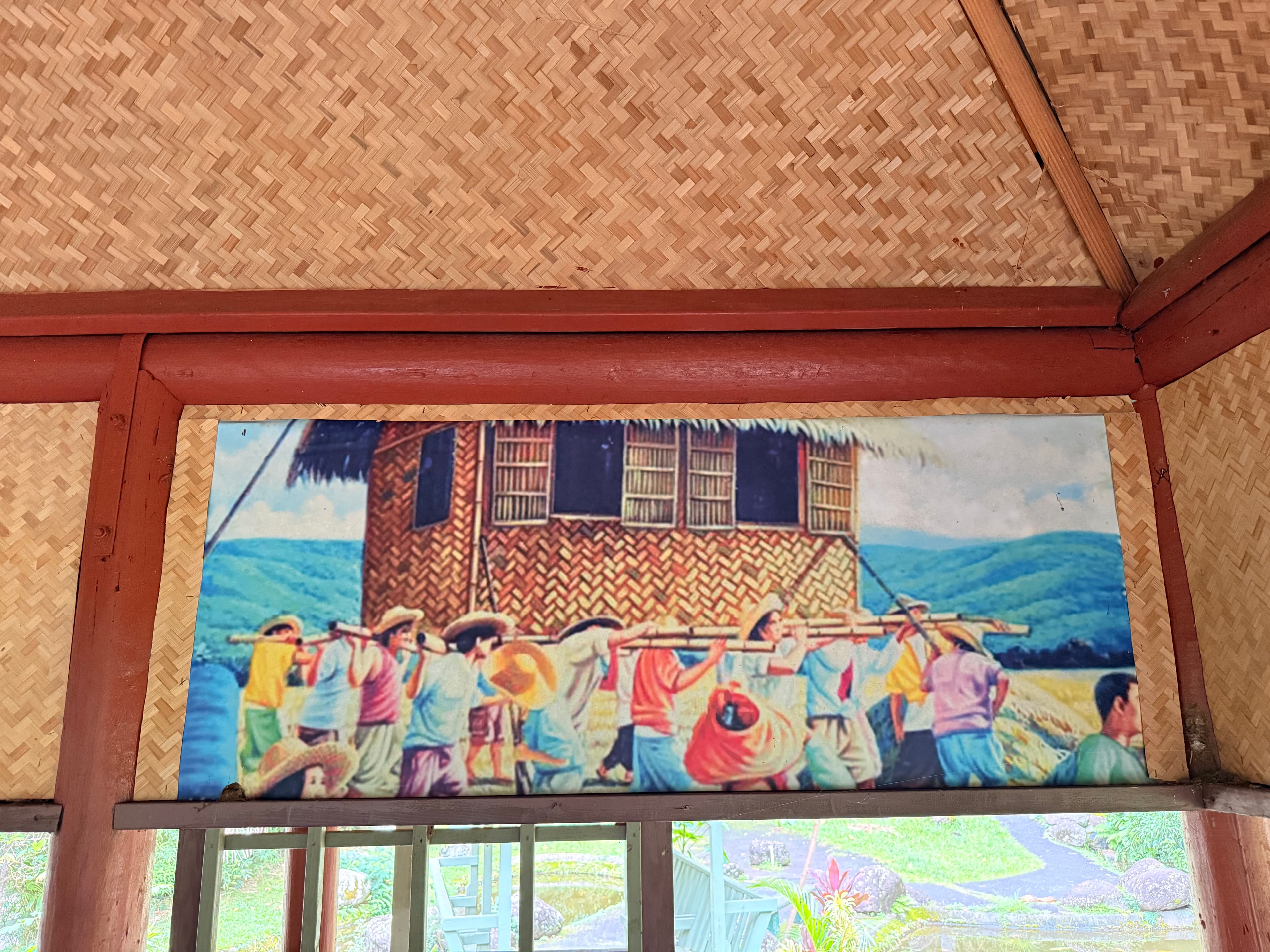 Painting of men in salakot hats carrying a bahay kubo on bamboo poles across green hills